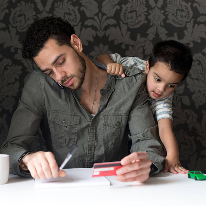 Father trying to focus on bills while his young son climbs on his shoulders, illustrating kids interrupting parents during conversations.