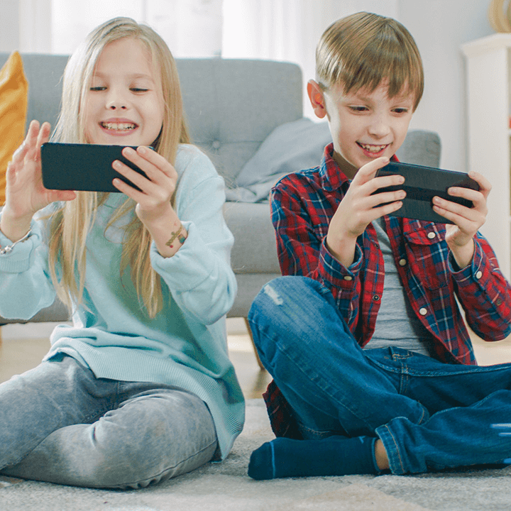 Two young children sitting on the floor at home, smiling and playing on smartphones—illustrating the challenge of kids spending too much time on screens.