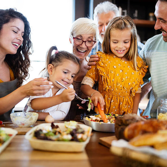 A family gathered in the kitchen preparing food together, smiling and enjoying a relaxed holiday moment with their children.