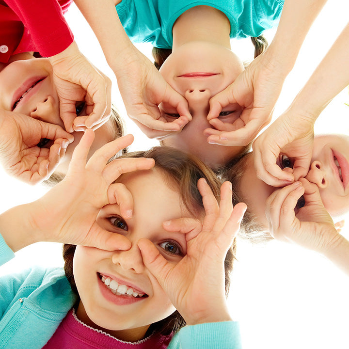 A group of smiling children lying in a circle and making playful hand-eye shapes, symbolizing joy, connection, and the positive behaviors kids notice from adults.